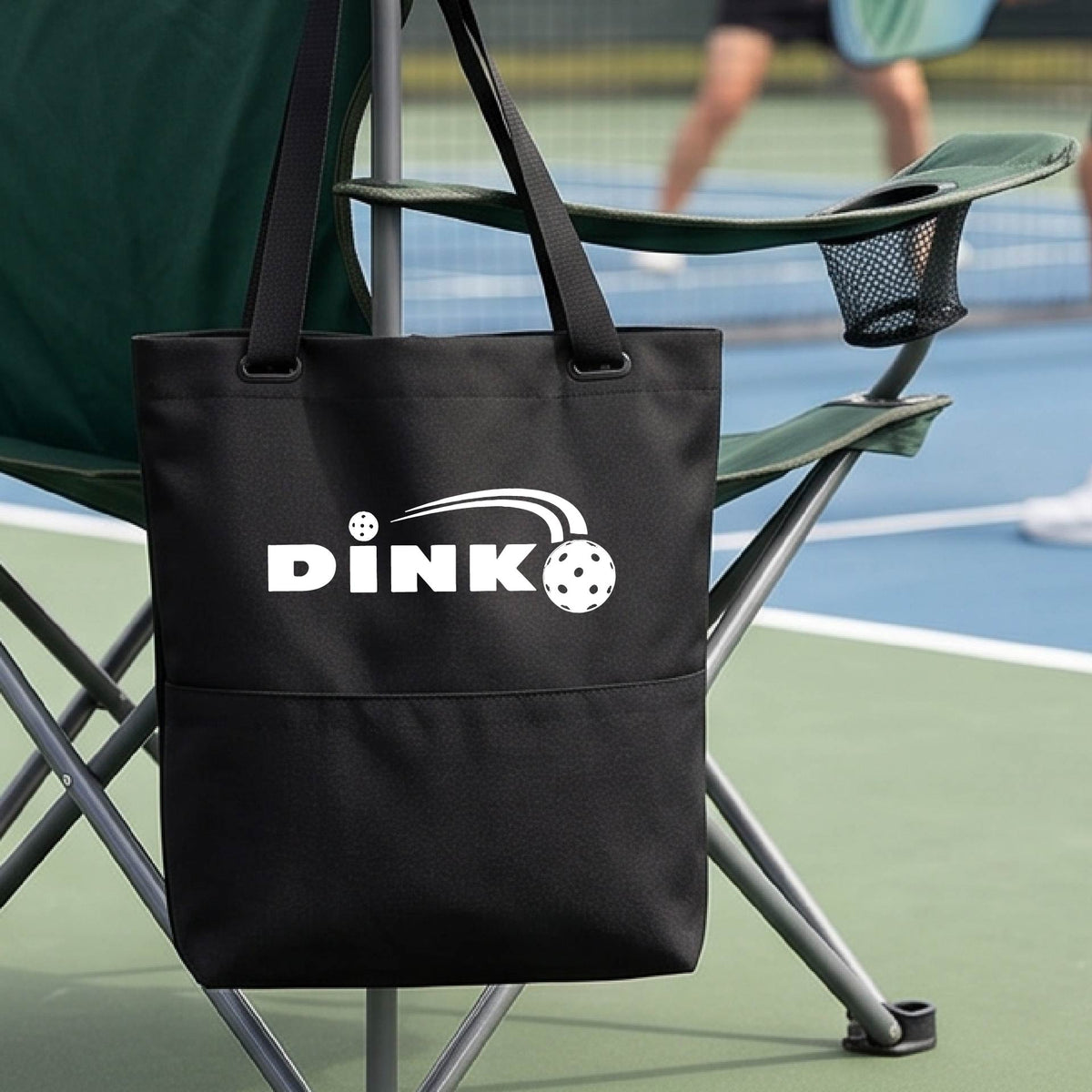 A black tote bag with a white 'DINK' logo with a white pickleball in flight hangs from a chair on a pickleball court. 