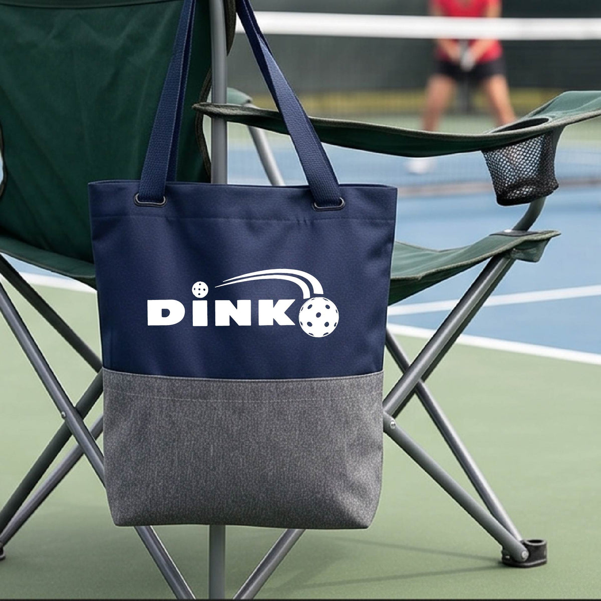 A navy blue and gray tote bag with a white 'DINK' logo with a white pickleball in flight hangs from a chair on a pickleball court. 