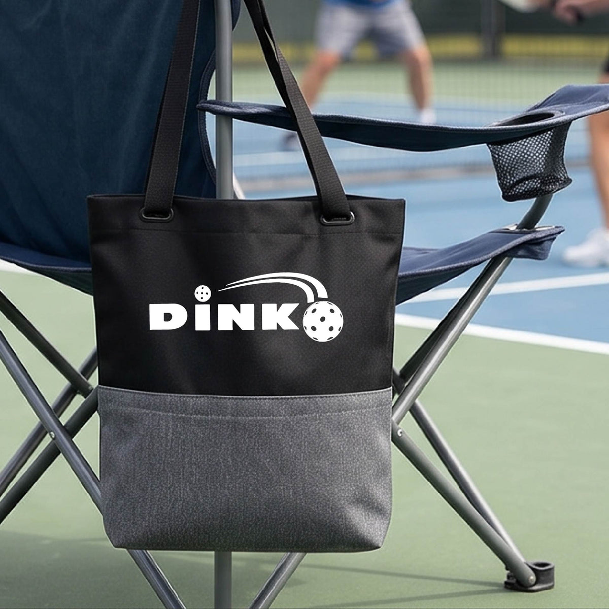 A black and gray tote bag with a white 'DINK' logo with a white pickleball in flight hangs from a chair on a pickleball court. 