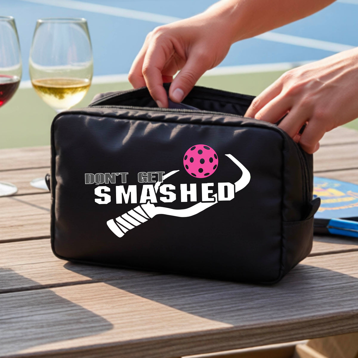 A black zippered cosmetic toiletry bag sits on a wooden table outdoors. The bag has a white graphic print that reads 'DON'T GET SMASHED' with a red pickelball and white pickleball paddle design.