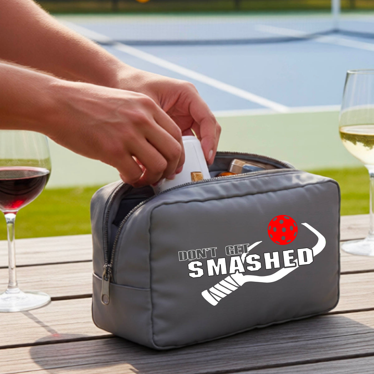 A gray zippered cosmetic toiletry bag sits on a wooden table outdoors. The bag has a white graphic print that reads 'DON'T GET SMASHED' with a red pickelball and white pickleball paddle design.