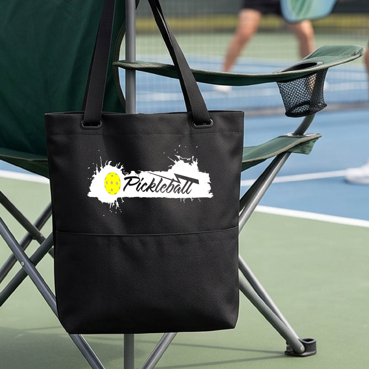 A black polyester tote bag with 'Pickleball' printed in white and a yellow pickleball splatter graphic, on a pickleball court.