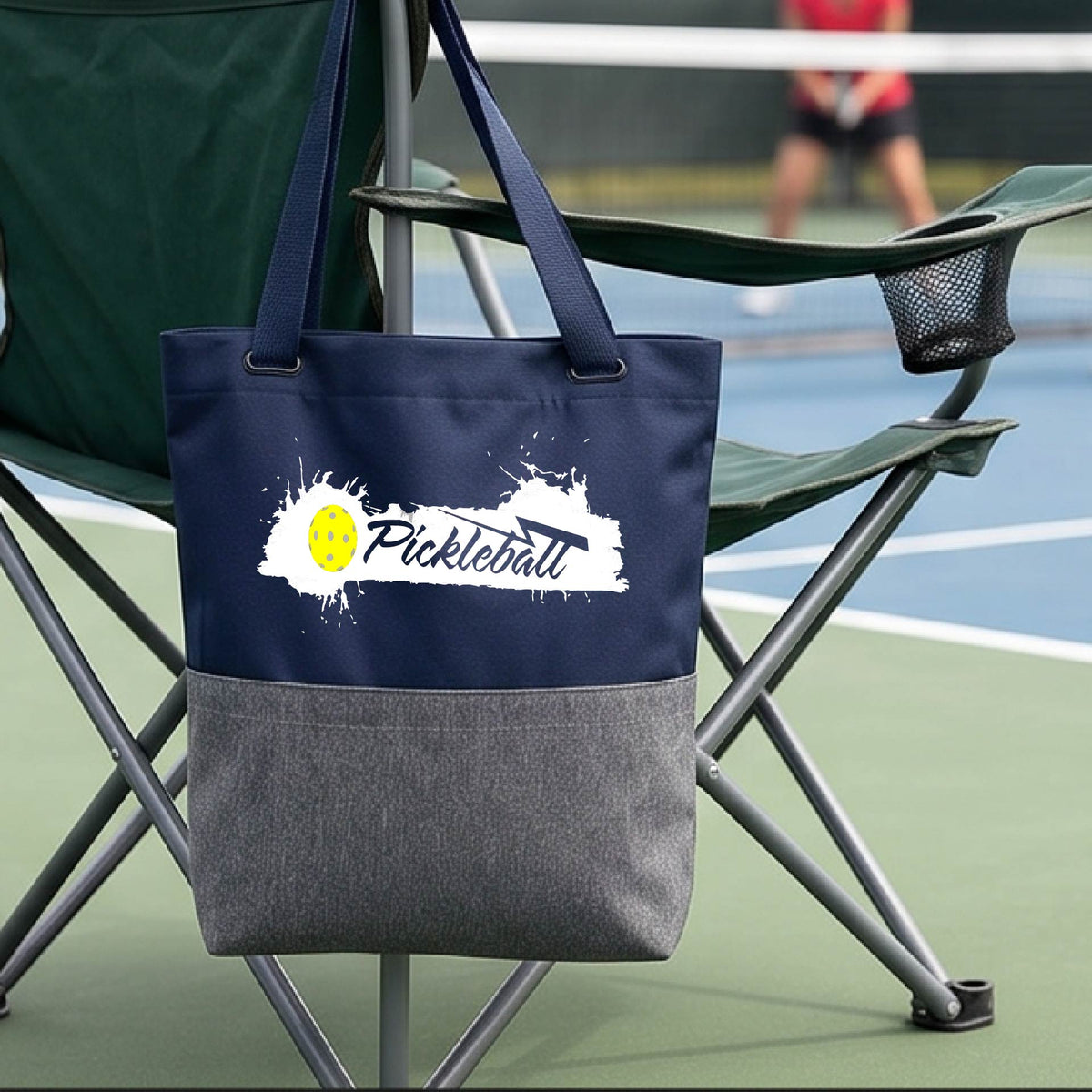 A navy blue and gray polyester tote bag with 'Pickleball' printed in white and a yellow pickleball splatter graphic, on a pickleball court.