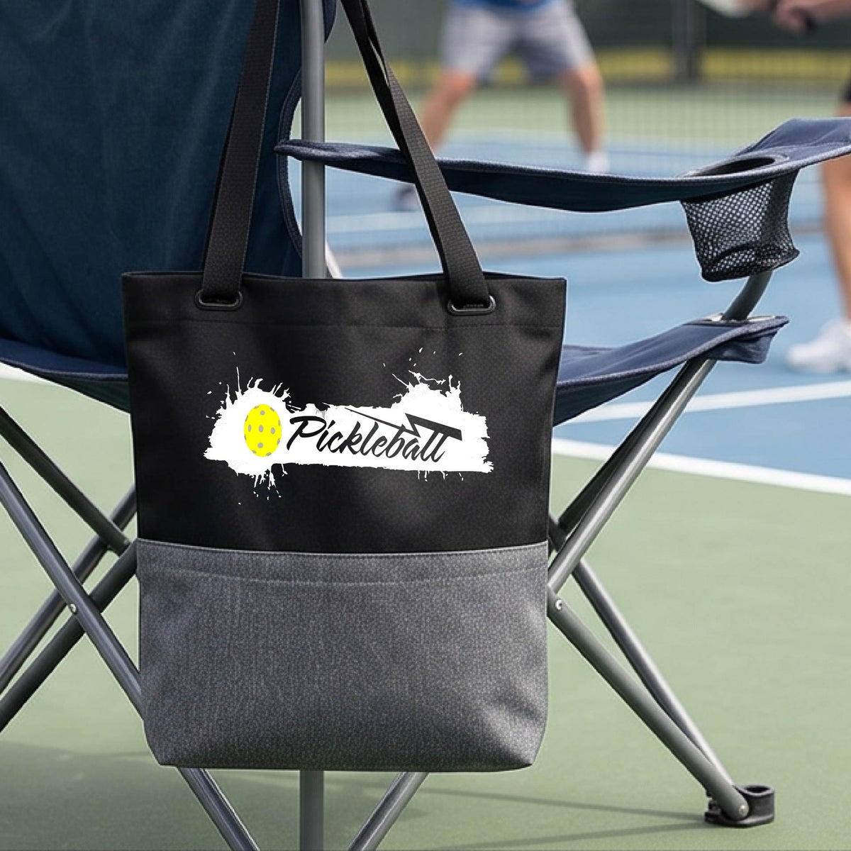A black and gray polyester tote bag with 'Pickleball' printed in white and a yellow pickleball splatter graphic, on a pickleball court.