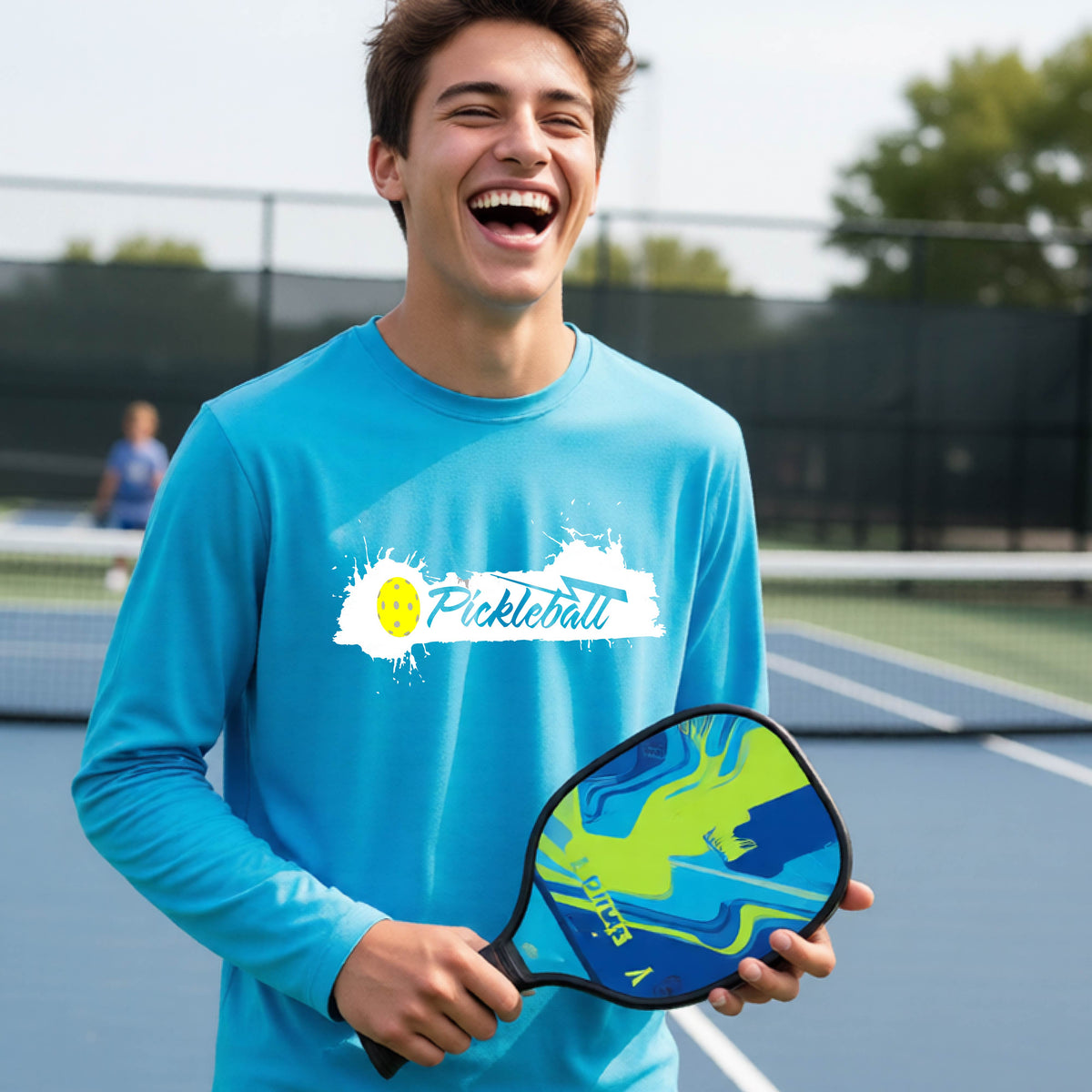A young person laughing on an outdoor tennis court wearing a bright blue long-sleeve t-shirt with a white and yellow 'Pickleball' splatter graphic on the chest.