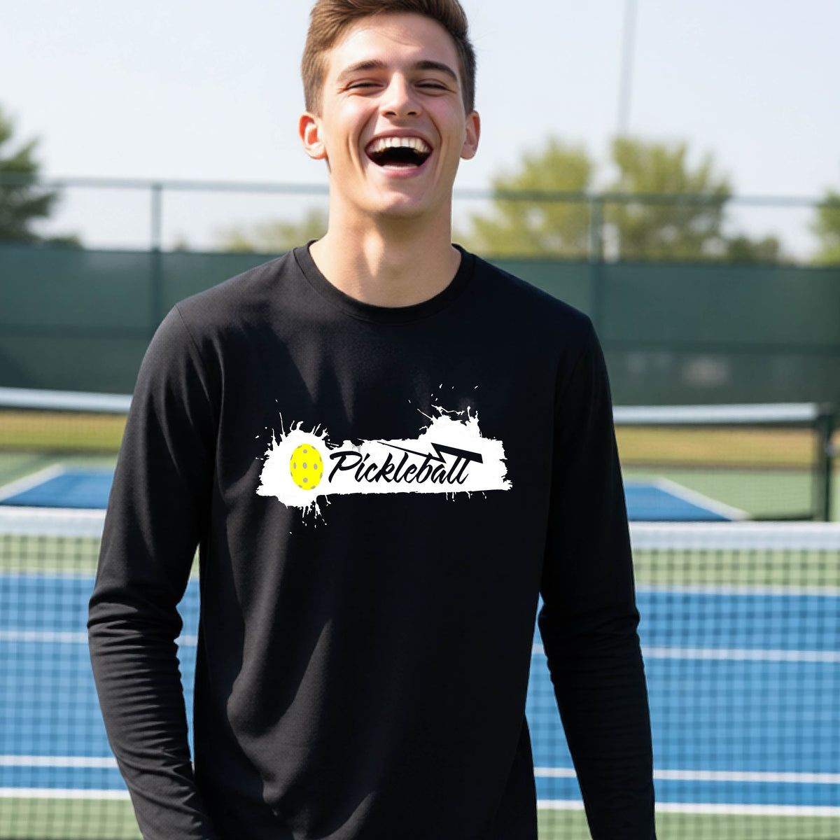 A young person laughing on an outdoor tennis court wearing a black long-sleeve t-shirt with a white and yellow 'Pickleball' splatter graphic on the chest.