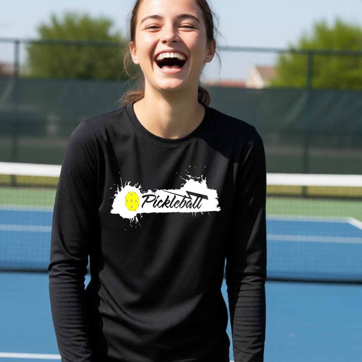 A young person laughing on an outdoor tennis court wearing a black long-sleeve t-shirt with a white and yellow 'Pickleball' splatter graphic on the chest.