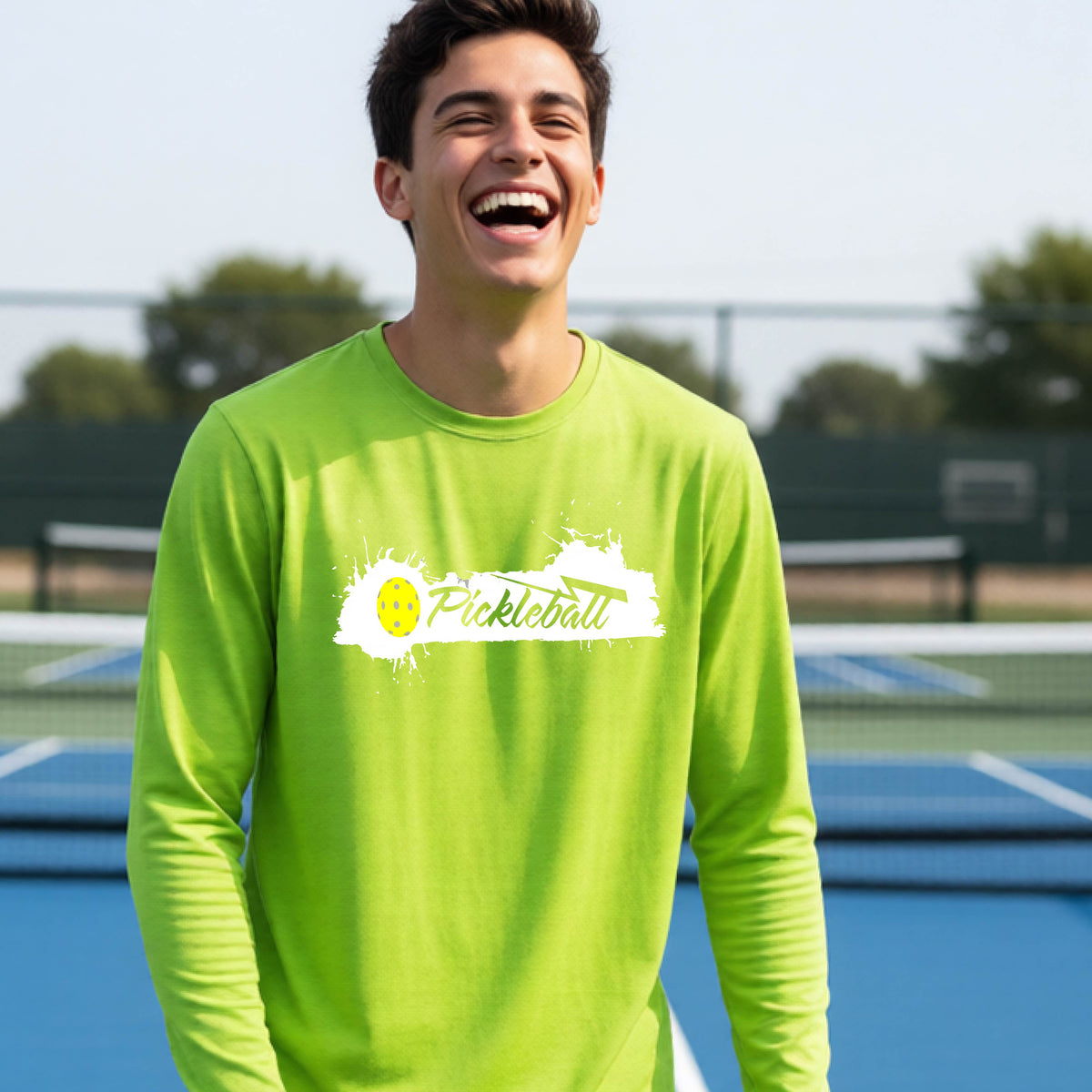 A young person laughing on an outdoor tennis court wearing a lime green long-sleeve t-shirt with a white and yellow 'Pickleball' splatter graphic on the chest.