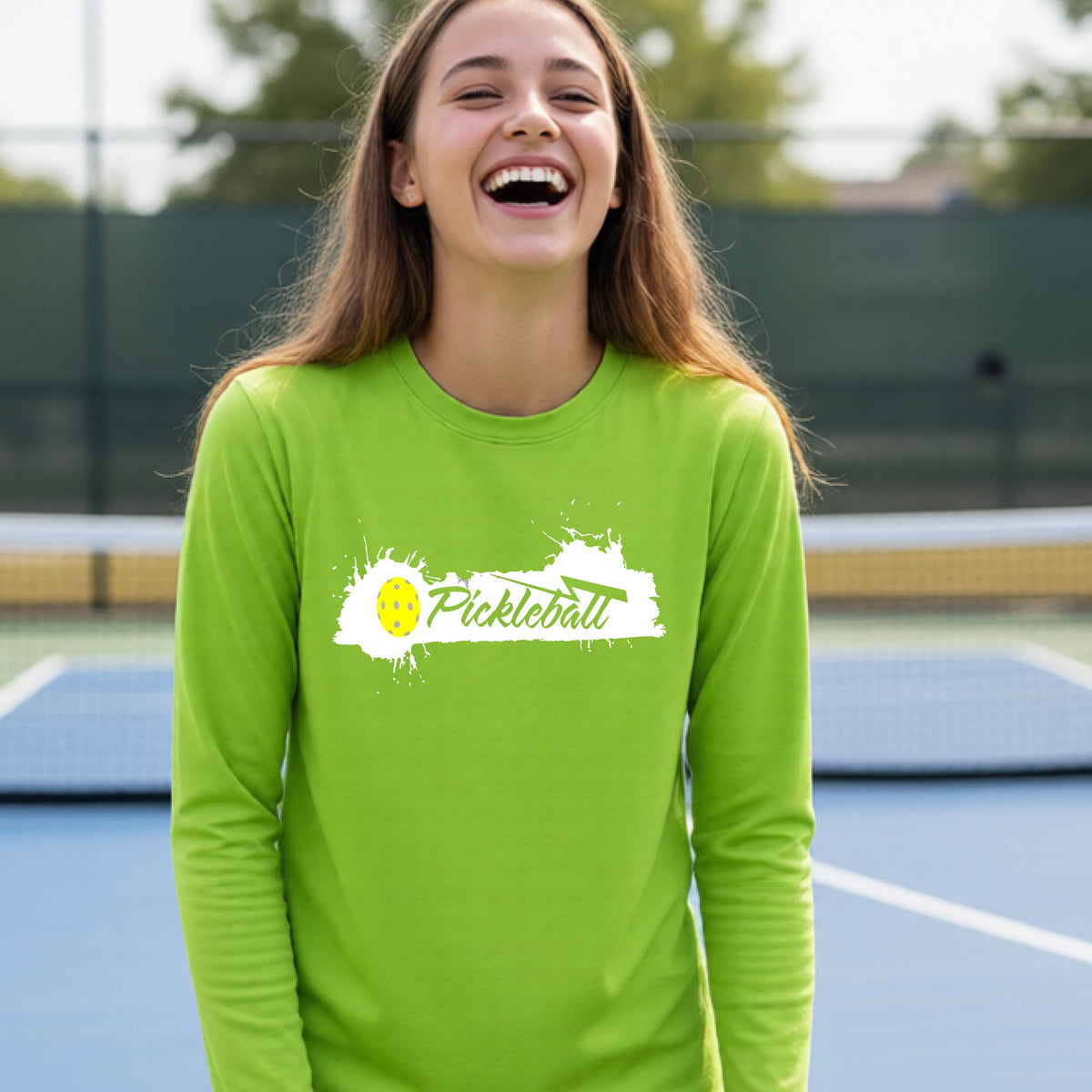 A young person laughing on an outdoor tennis court wearing a lime green long-sleeve t-shirt with a white and yellow 'Pickleball' splatter graphic on the chest.