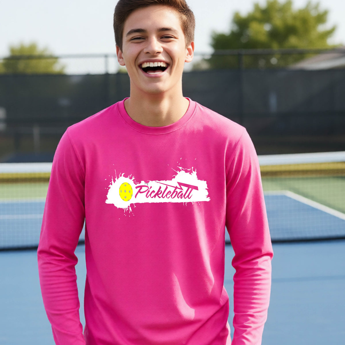 A young person laughing on an outdoor tennis court wearing a bright pink long-sleeve t-shirt with a white and yellow 'Pickleball' splatter graphic on the chest.