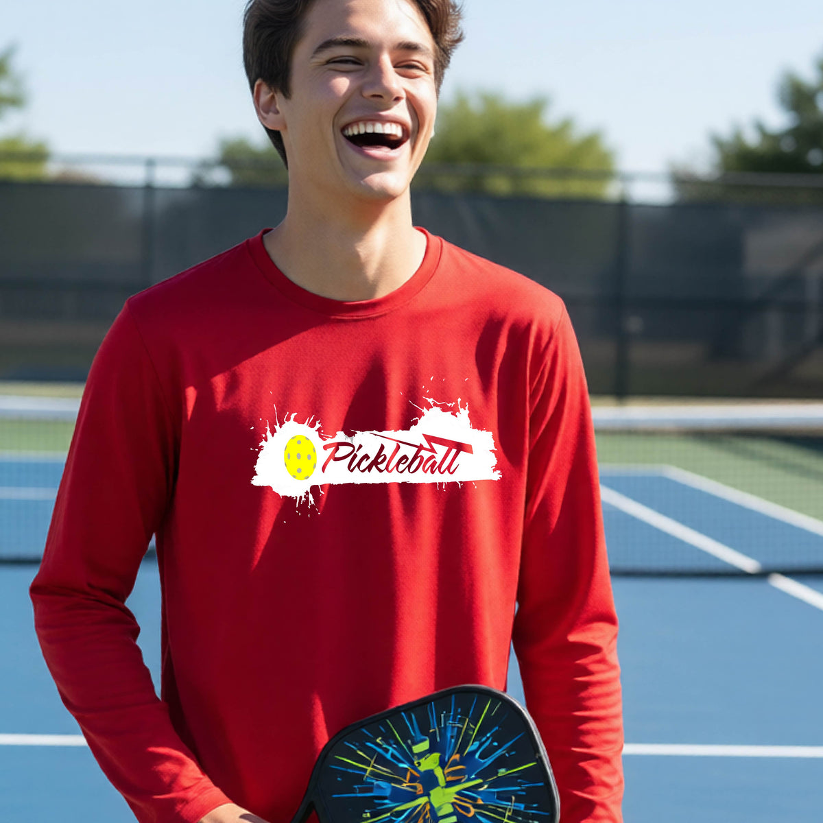 A young person laughing on an outdoor tennis court wearing a red long-sleeve t-shirt with a white and yellow 'Pickleball' splatter graphic on the chest.