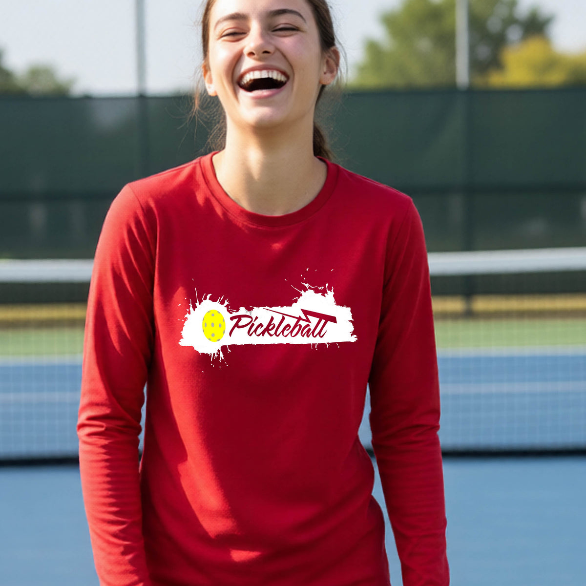 A young person laughing on an outdoor tennis court wearing a red long-sleeve t-shirt with a white and yellow 'Pickleball' splatter graphic on the chest.
