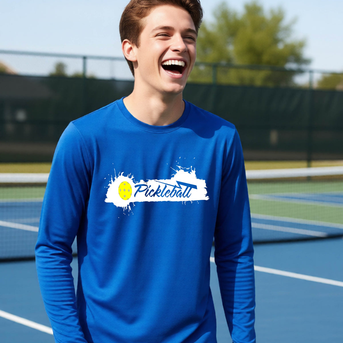 A young person laughing on an outdoor tennis court wearing a royal blue long-sleeve t-shirt with a white and yellow 'Pickleball' splatter graphic on the chest.