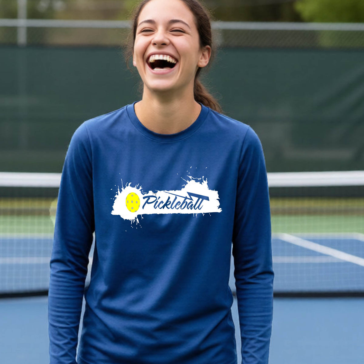 A young person laughing on an outdoor tennis court wearing a royal blue long-sleeve t-shirt with a white and yellow 'Pickleball' splatter graphic on the chest.