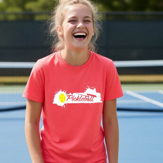 A young person laughing on an outdoor pickleball court, wearing a coral pink t-shirt with a white and yellow 'Pickleball' splatter graphic on the chest.