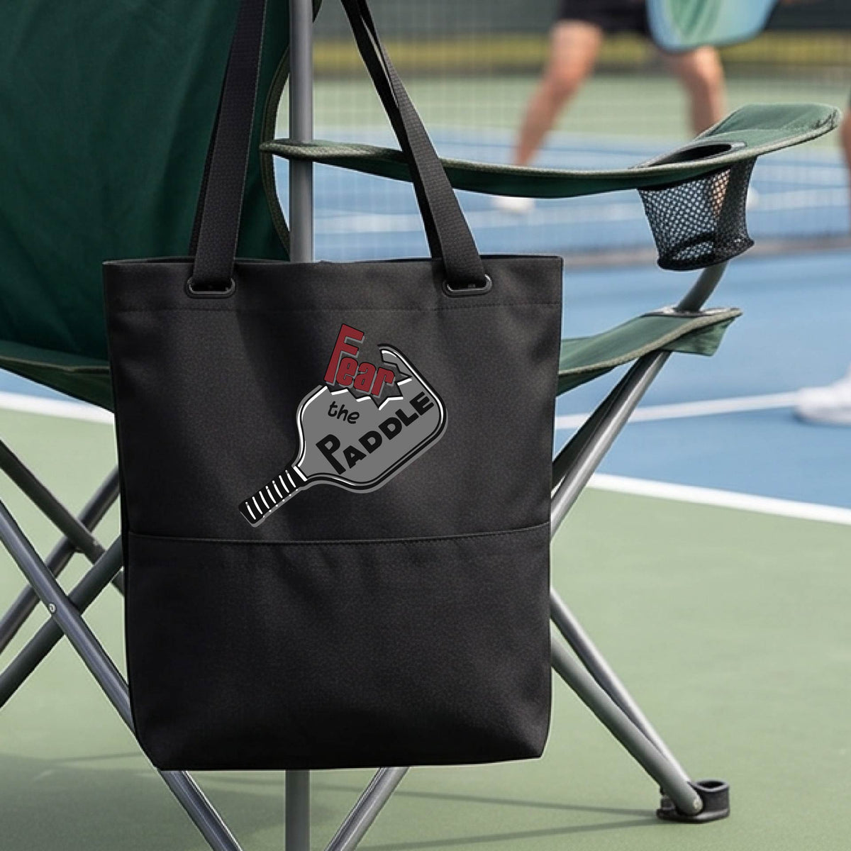 A black tote bag with a pickleball graphic of the text 'FEAR THE PADDLE' cut out into a pickleball paddle, with a pickleball court in the background. 