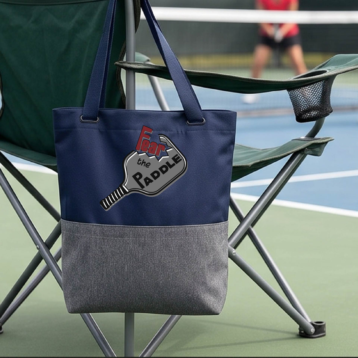 A blue and gray tote bag with a pickleball graphic of the text 'FEAR THE PADDLE' cut out into a pickleball paddle, with a pickleball court in the background. 