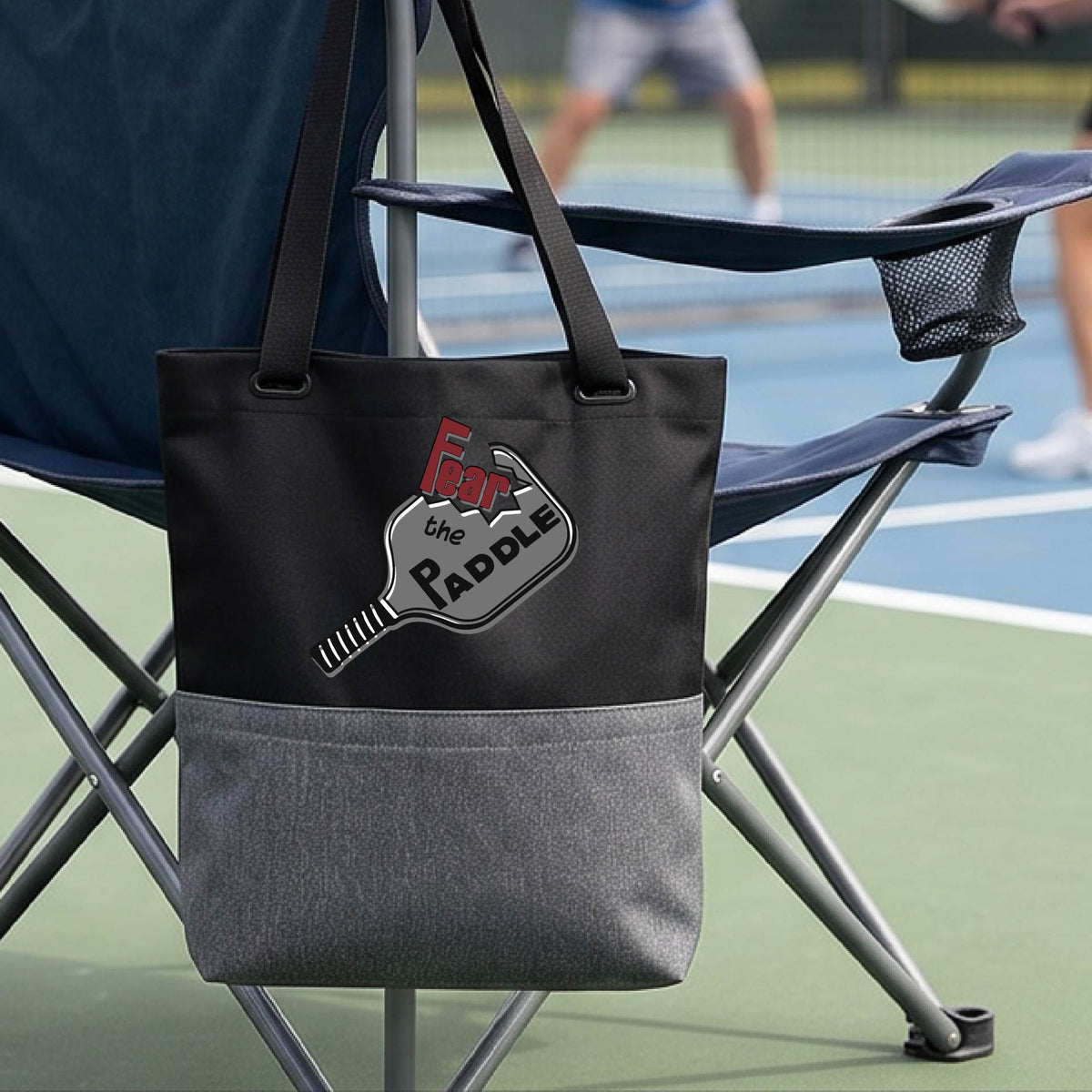 A black and gray tote bag with a pickleball graphic of the text 'FEAR THE PADDLE' cut out into a pickleball paddle, with a pickleball court in the background. 