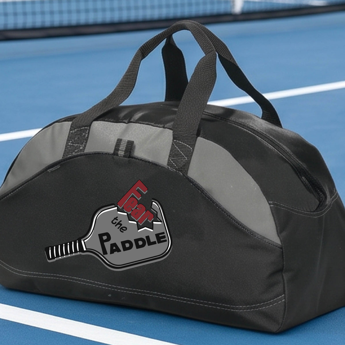 A gray and black duffel bag with a pickleball graphic of the text 'FEAR THE PADDLE' cut into a pickleball paddle, set on a background of a pickleball court. 