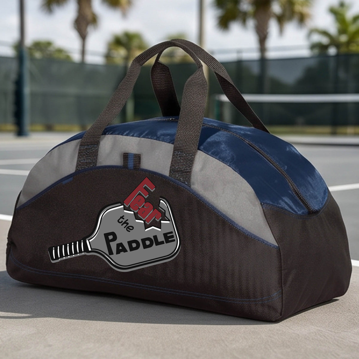 A navy blue, gray and black duffel bag with a pickleball graphic of the text 'FEAR THE PADDLE' cut into a pickleball paddle, set on a background of a pickleball court. 