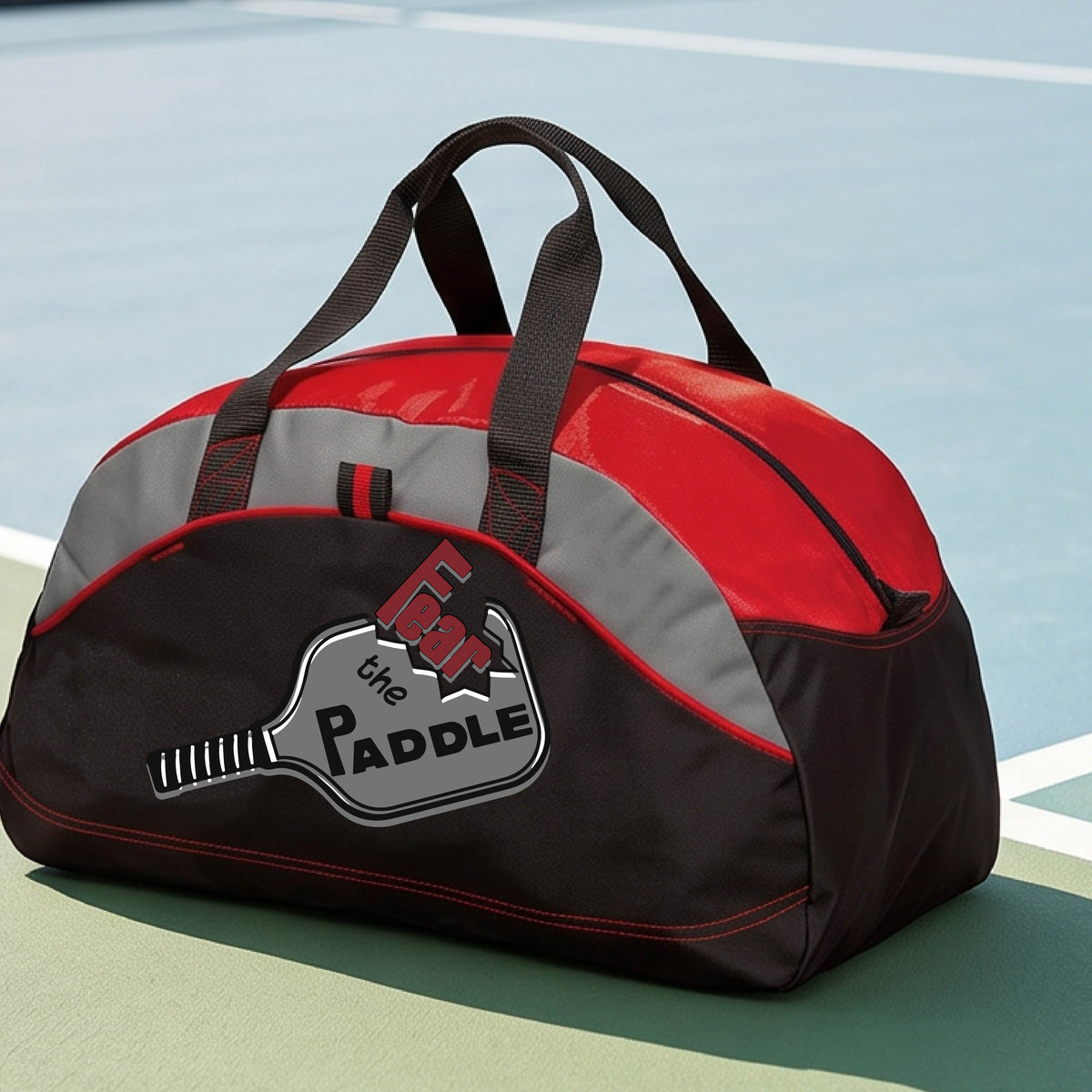 A red, gray and black duffel bag with a pickleball graphic of the text 'FEAR THE PADDLE' cut into a pickleball paddle, set on a background of a pickleball court. 