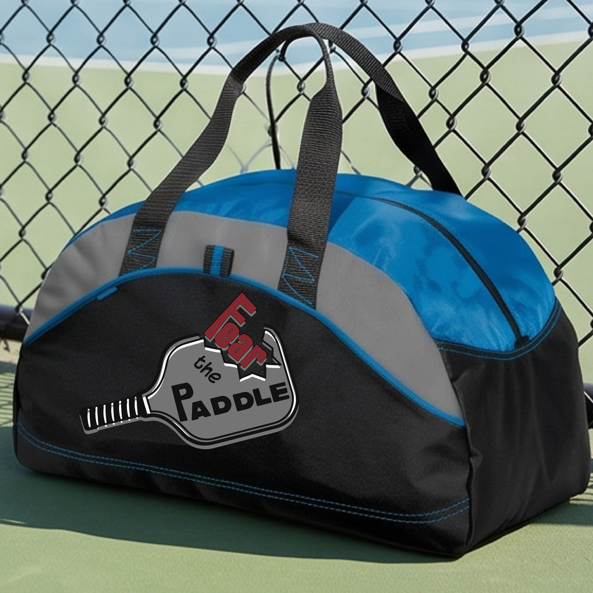 A royal blue, gray and black duffel bag with a pickleball graphic of the text 'FEAR THE PADDLE' cut into a pickleball paddle, set on a background of a pickleball court. 