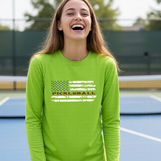 A young person wearing a lime green long-sleeve t-shirt with a pickleball graphic of a distressed flag made out of pickleballs and the text 'PICKLEBALL' replacing one of the stripes, standing on a pickleball court.