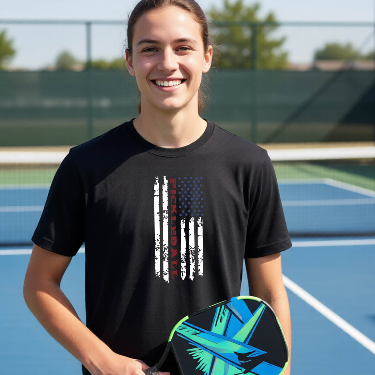 A young person wearing a black short-sleeve  t-shirt with a pickleball graphic of a distressed flag made out of pickleballs and the text 'PICKLEBALL' replacing one of the stripes, standing on a pickleball court.