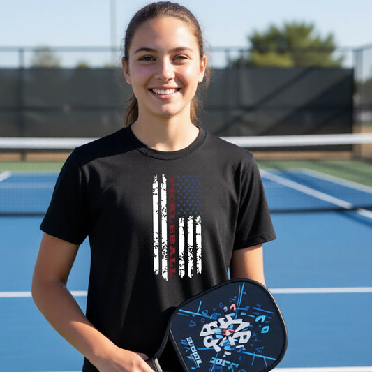 A young person wearing a black short-sleeve  t-shirt with a pickleball graphic of a distressed flag made out of pickleballs and the text 'PICKLEBALL' replacing one of the stripes, standing on a pickleball court.