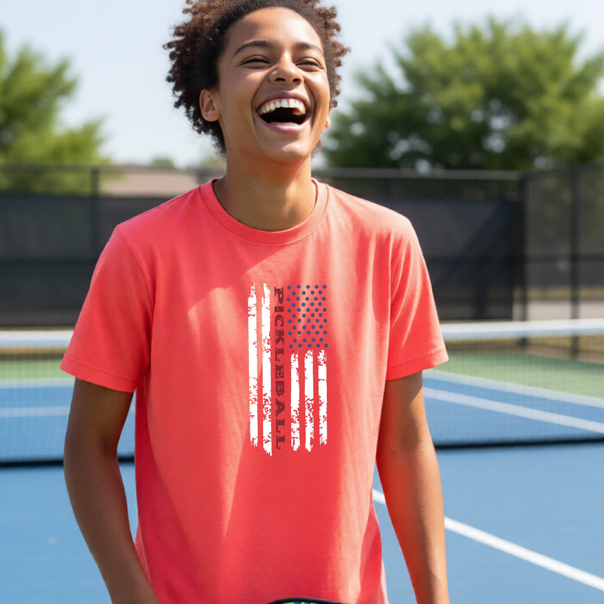 A young person wearing a coral pink short-sleeve  t-shirt with a pickleball graphic of a distressed flag made out of pickleballs and the text 'PICKLEBALL' replacing one of the stripes, standing on a pickleball court.