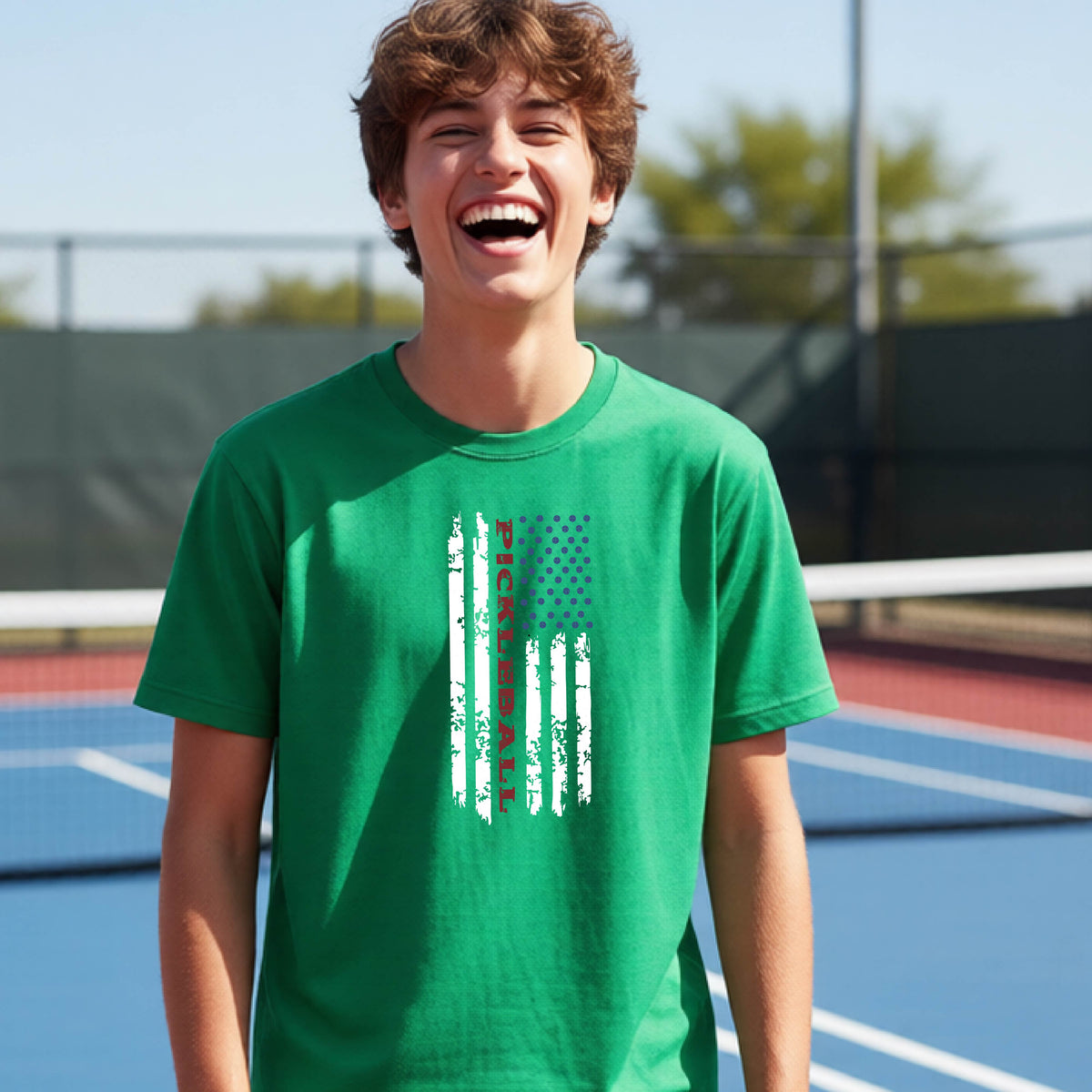 A young person wearing a green short-sleeve  t-shirt with a pickleball graphic of a distressed flag made out of pickleballs and the text 'PICKLEBALL' replacing one of the stripes, standing on a pickleball court.
