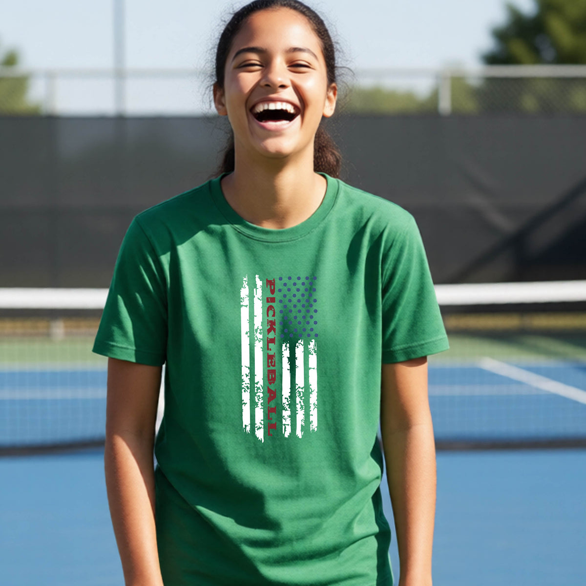 A young person wearing a green short-sleeve  t-shirt with a pickleball graphic of a distressed flag made out of pickleballs and the text 'PICKLEBALL' replacing one of the stripes, standing on a pickleball court.