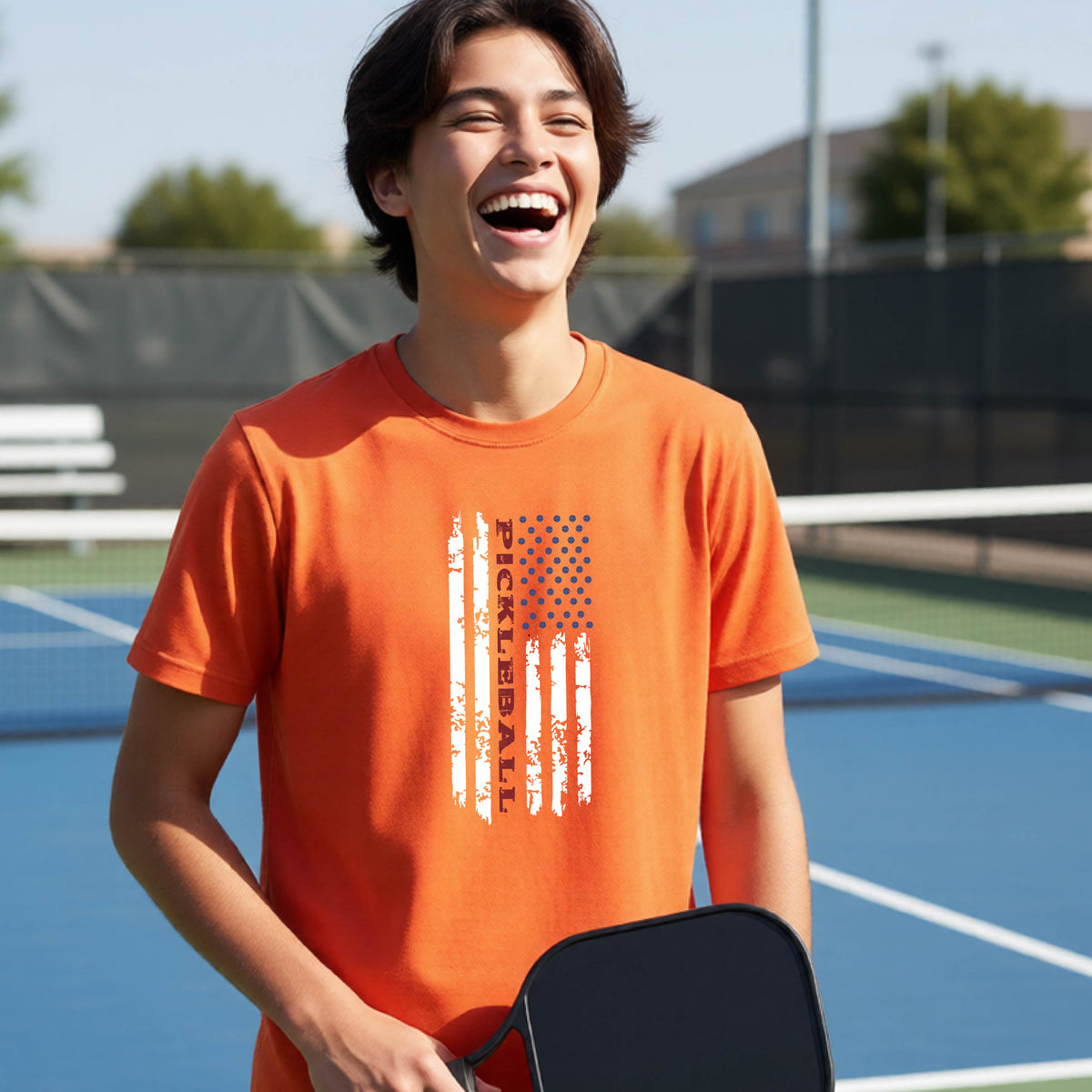 A young person wearing an orange short-sleeve  t-shirt with a pickleball graphic of a distressed flag made out of pickleballs and the text 'PICKLEBALL' replacing one of the stripes, standing on a pickleball court.