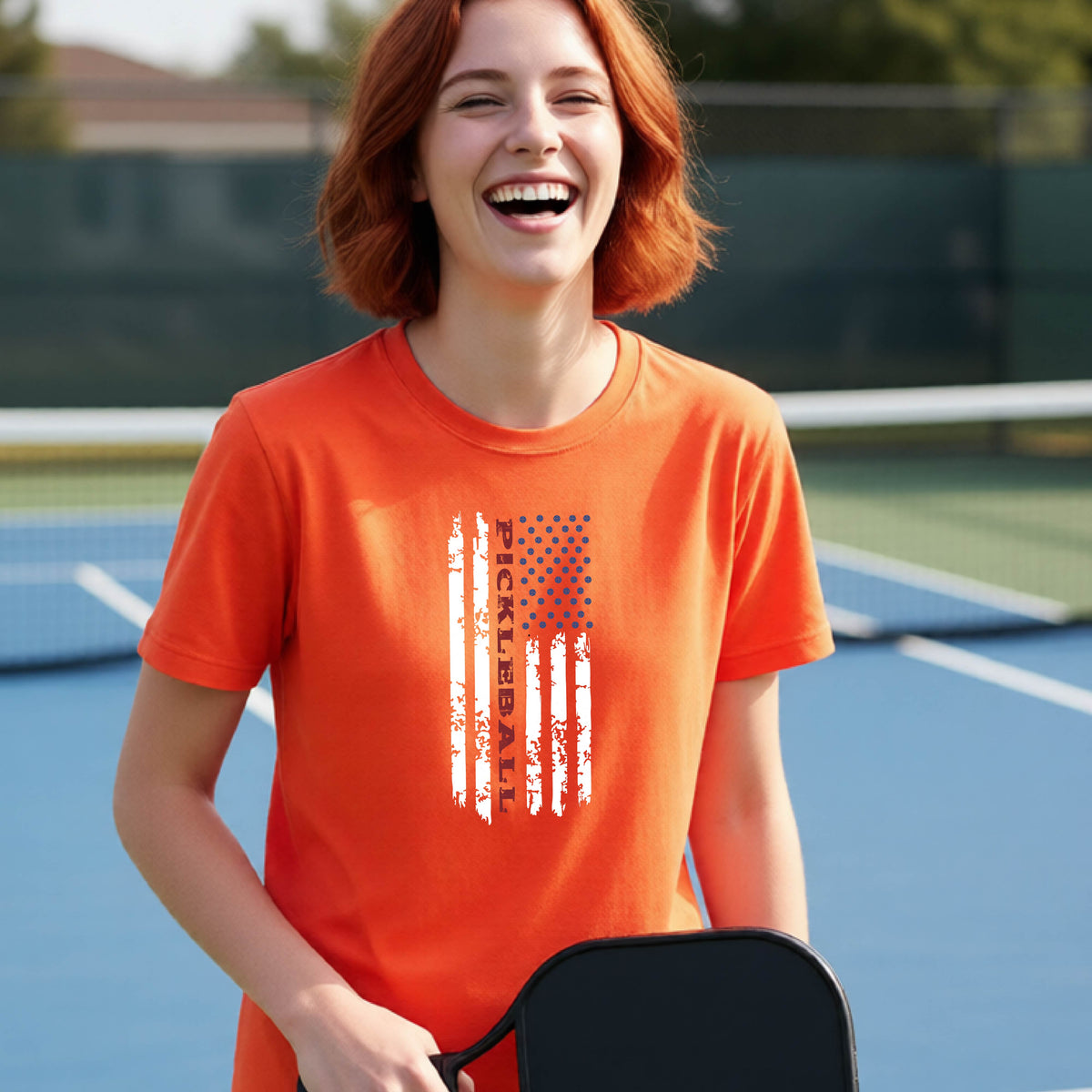 A young person wearing an orange short-sleeve  t-shirt with a pickleball graphic of a distressed flag made out of pickleballs and the text 'PICKLEBALL' replacing one of the stripes, standing on a pickleball court.
