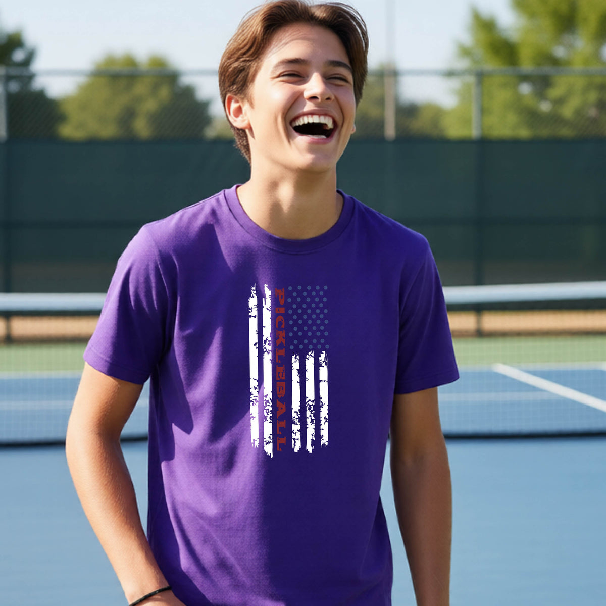 A young person wearing a purple short-sleeve  t-shirt with a pickleball graphic of a distressed flag made out of pickleballs and the text 'PICKLEBALL' replacing one of the stripes, standing on a pickleball court.