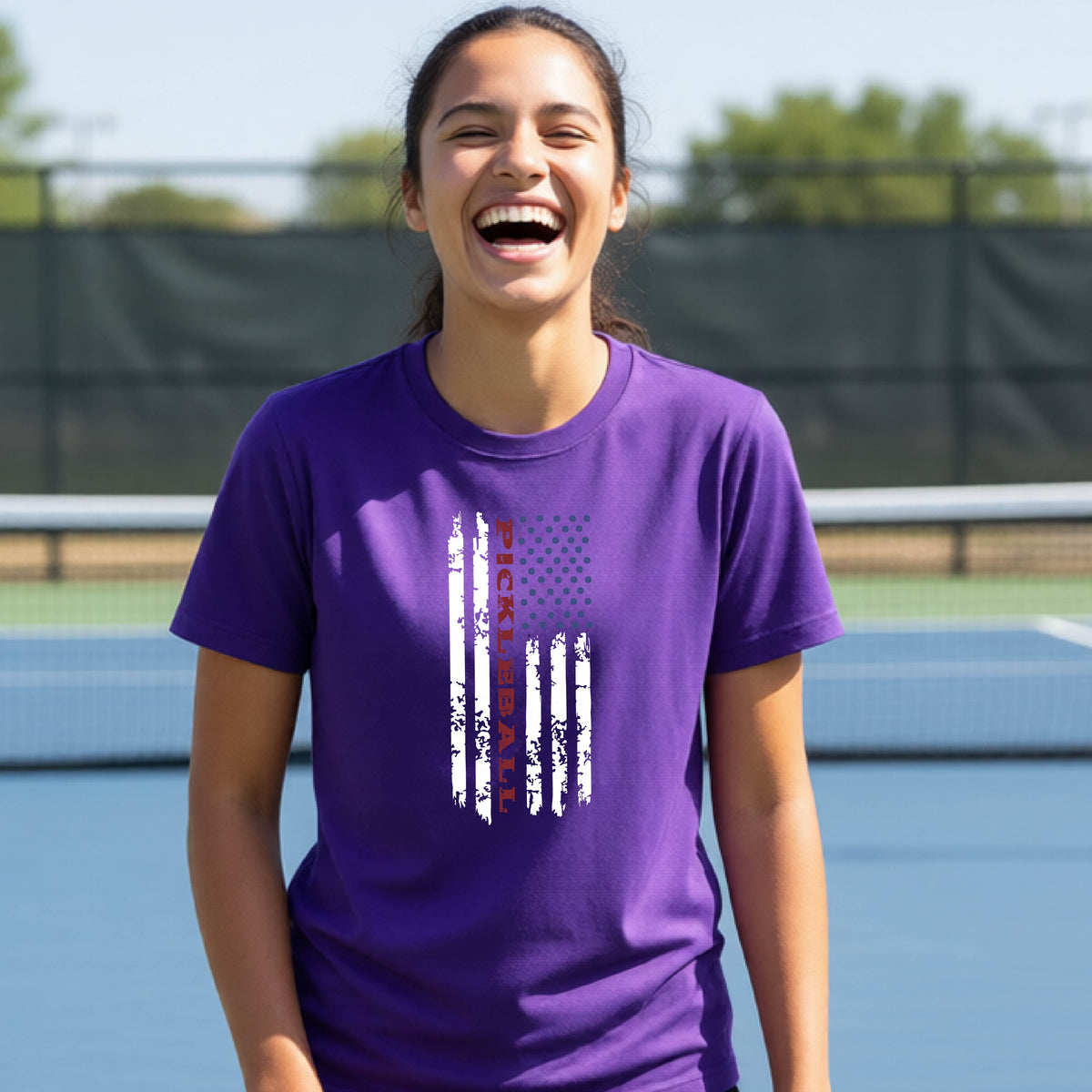 A young person wearing a purple short-sleeve  t-shirt with a pickleball graphic of a distressed flag made out of pickleballs and the text 'PICKLEBALL' replacing one of the stripes, standing on a pickleball court.