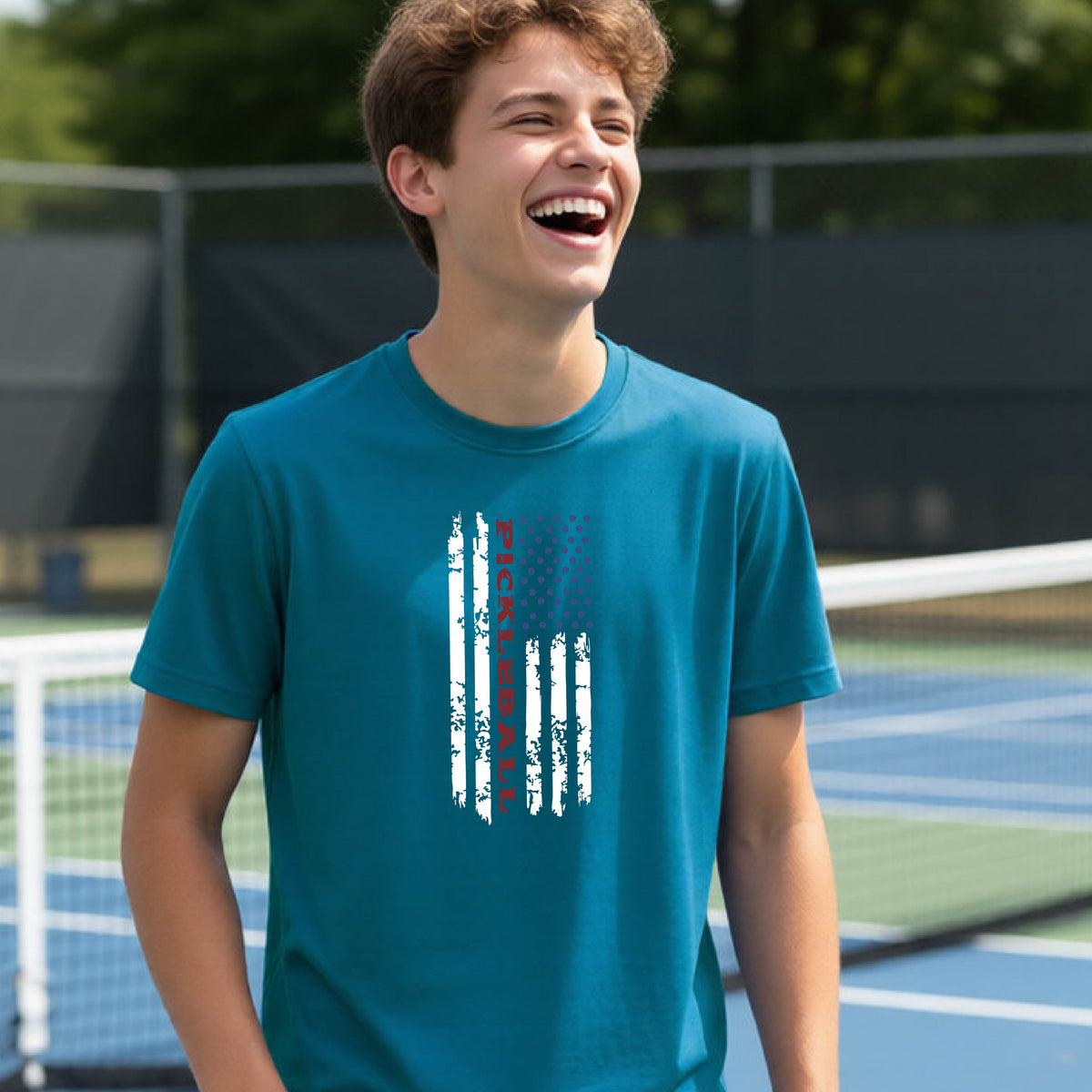 A young person wearing a teal short-sleeve  t-shirt with a pickleball graphic of a distressed flag made out of pickleballs and the text 'PICKLEBALL' replacing one of the stripes, standing on a pickleball court.
