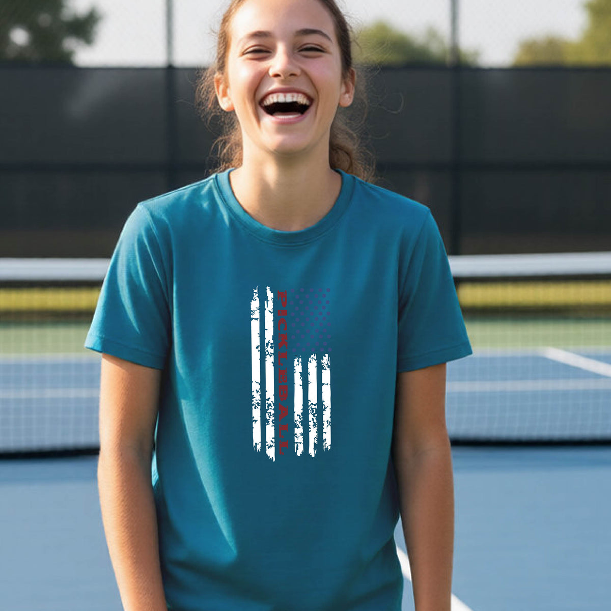 A young person wearing a teal short-sleeve  t-shirt with a pickleball graphic of a distressed flag made out of pickleballs and the text 'PICKLEBALL' replacing one of the stripes, standing on a pickleball court.