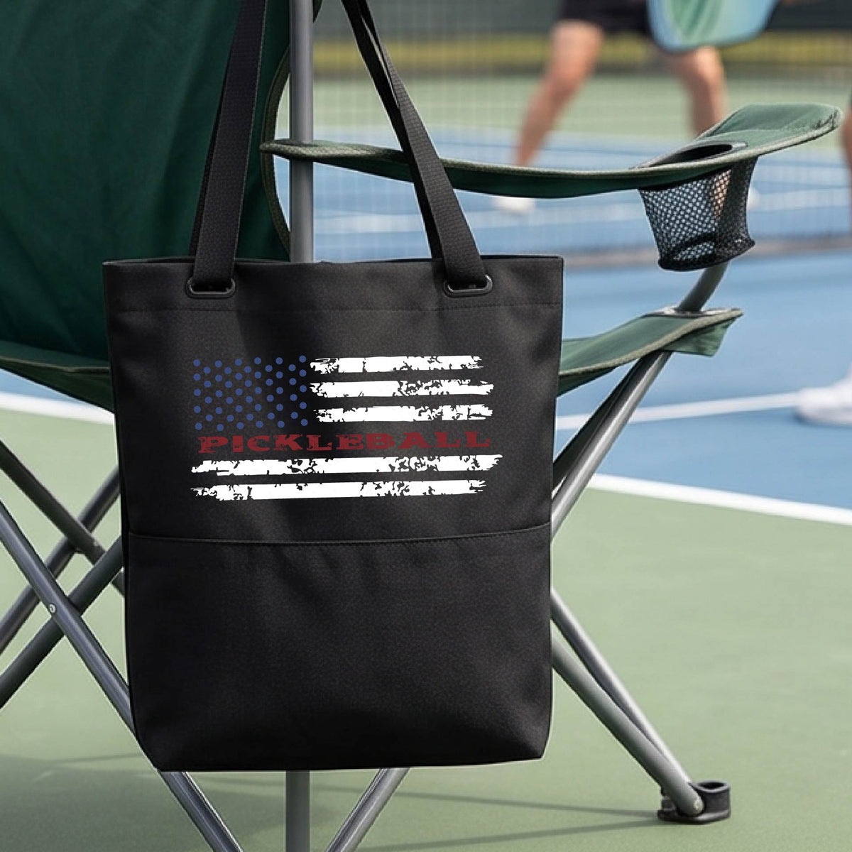 A black  tote bag with a pickleball graphic of a distressed flag made out of pickleballs and the text 'PICKLEBALL' replacing one of the stripes, standing on a pickleball court.