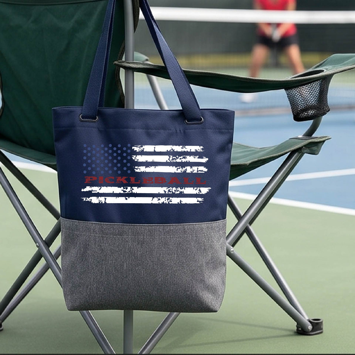 A blue and gray tote bag with a pickleball graphic of a distressed flag made out of pickleballs and the text 'PICKLEBALL' replacing one of the stripes, standing on a pickleball court.