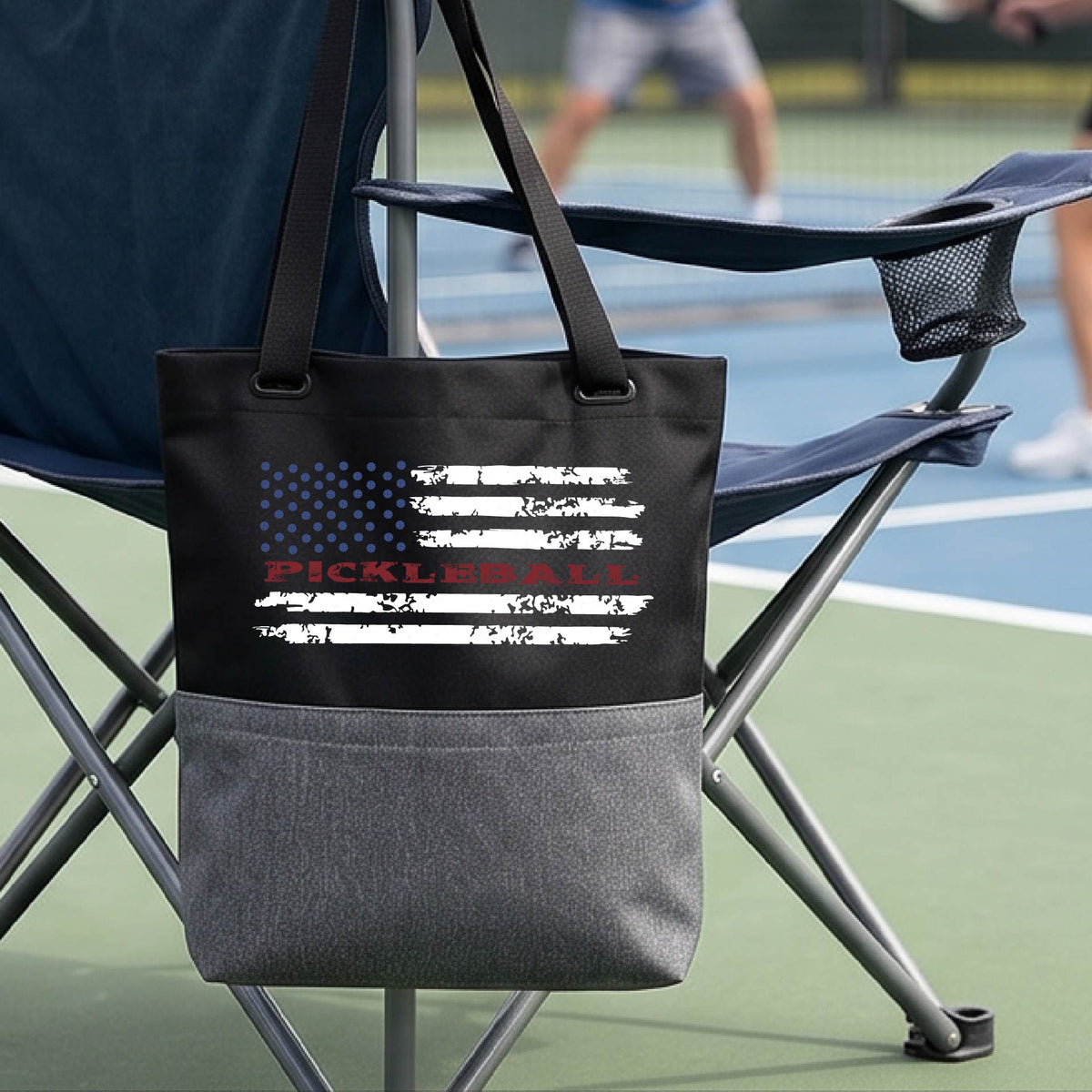 A black and gray tote bag with a pickleball graphic of a distressed flag made out of pickleballs and the text 'PICKLEBALL' replacing one of the stripes, standing on a pickleball court.