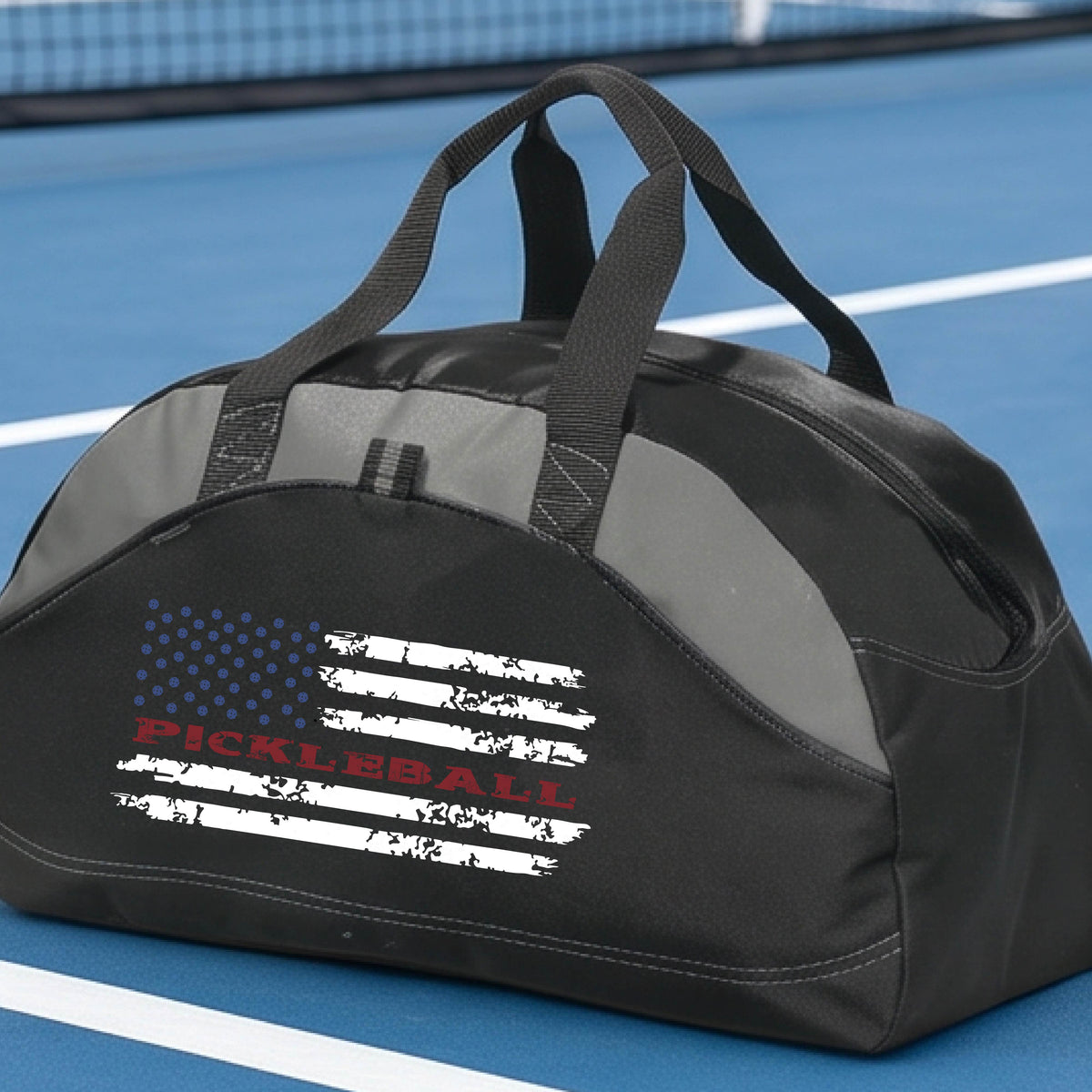 A black and gray duffel bag with a pickleball graphic of a distressed flag made out of pickleballs and the text 'PICKLEBALL' replacing one of the stripes, standing on a pickleball court.