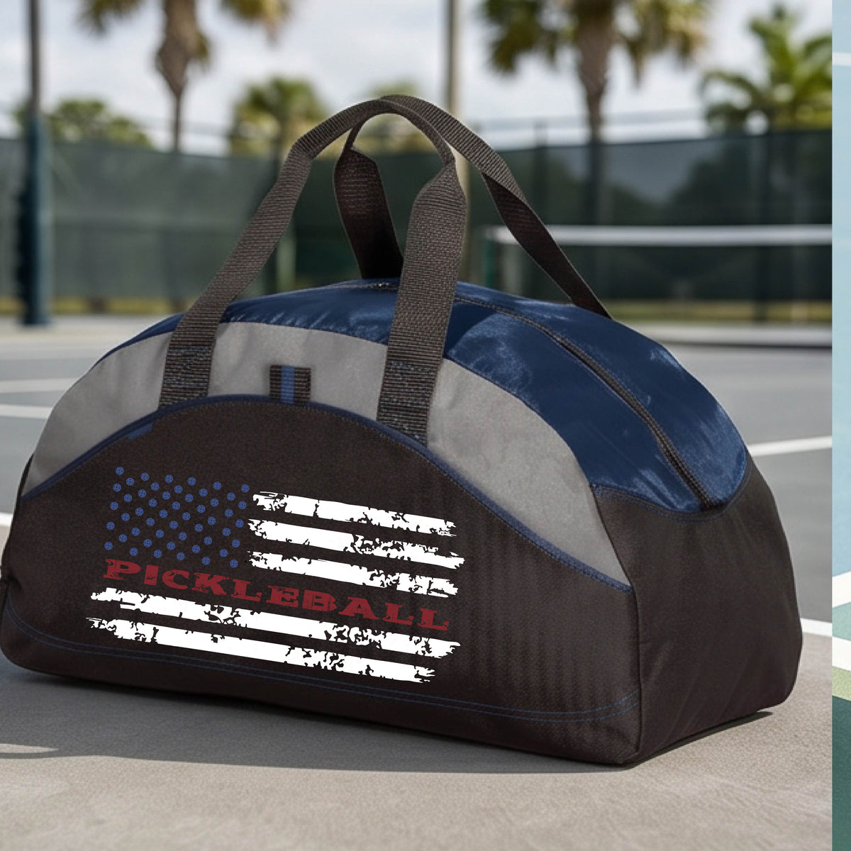 A navy blue, black and gray duffel bag with a pickleball graphic of a distressed flag made out of pickleballs and the text 'PICKLEBALL' replacing one of the stripes, standing on a pickleball court.