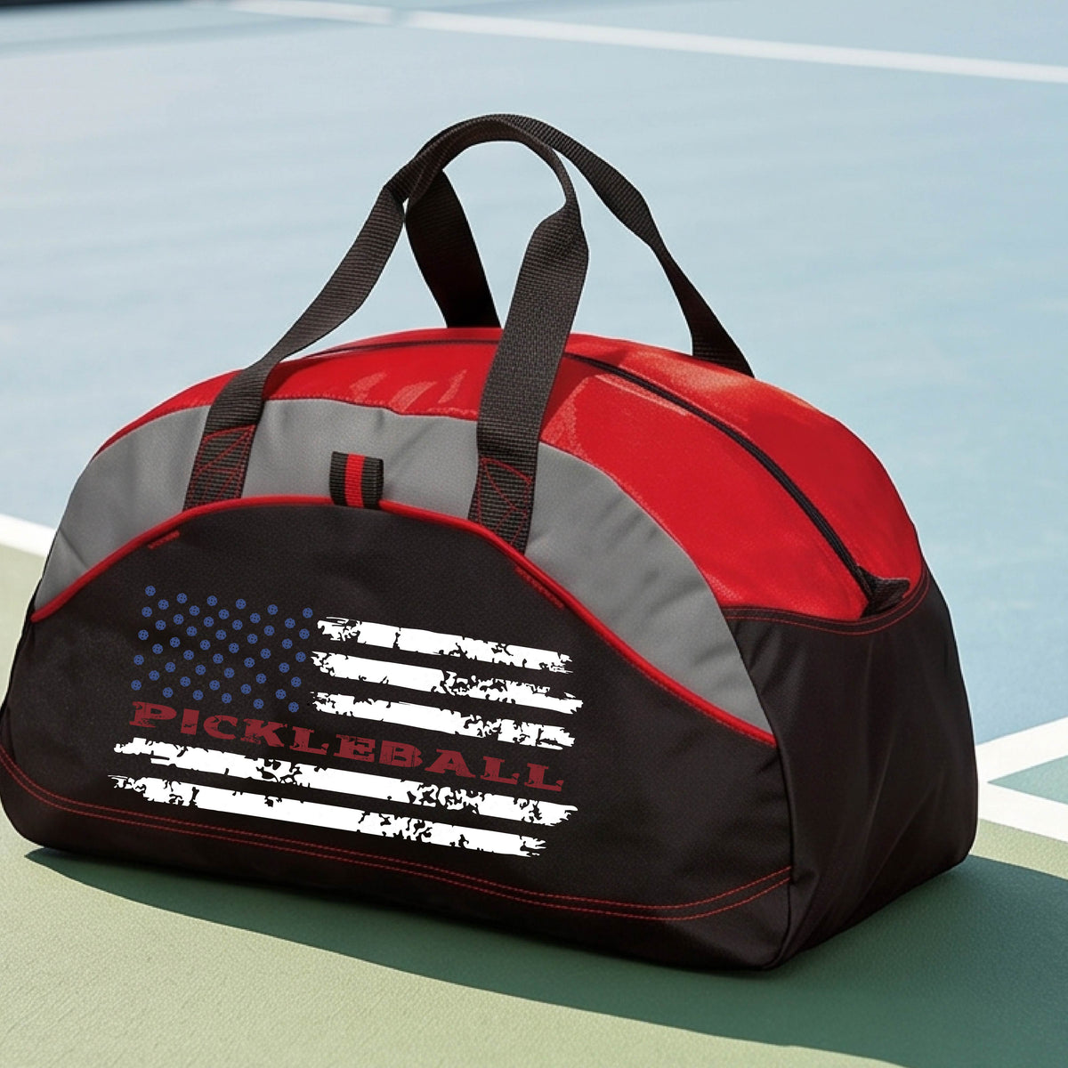A red, black and gray duffel bag with a pickleball graphic of a distressed flag made out of pickleballs and the text 'PICKLEBALL' replacing one of the stripes, standing on a pickleball court.