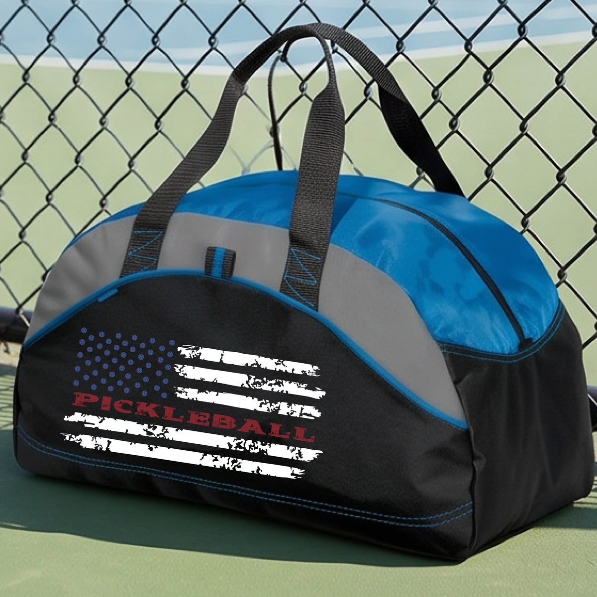 A royal blue, black and gray duffel bag with a pickleball graphic of a distressed flag made out of pickleballs and the text 'PICKLEBALL' replacing one of the stripes, standing on a pickleball court.