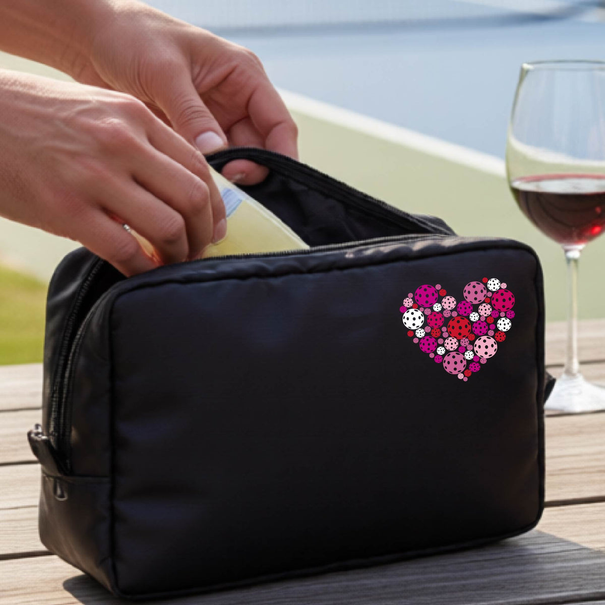A black zippered toiletry bag with a heart-shaped pattern in pink, purple, and white, pickleballs shown on a wooden table with a wine glass and a person's hands in the background.