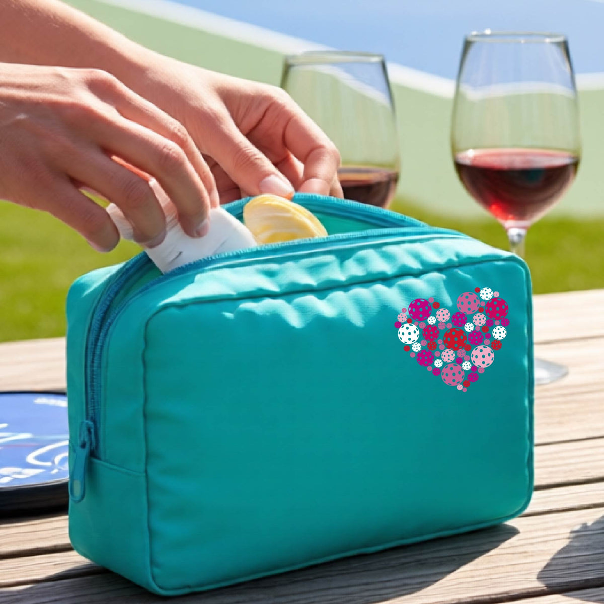 A turquoise zippered toiletry bag with a heart-shaped pattern in pink, purple, and white, pickleballs shown on a wooden table with a wine glass and a person's hands in the background.