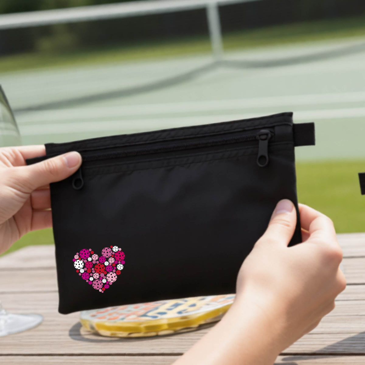 A black zippered toiletry bag with a heart-shaped pattern in pink, purple, and white, pickleballs shown on a wooden table with a wine glass and a person's hands in the background.