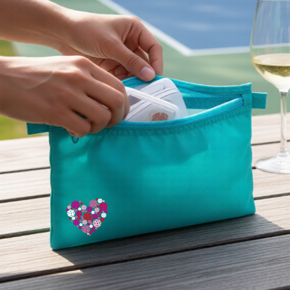 A turquoise zippered toiletry bag with a heart-shaped pattern in pink, purple, and white, pickleballs shown on a wooden table with a wine glass and a person's hands in the background.