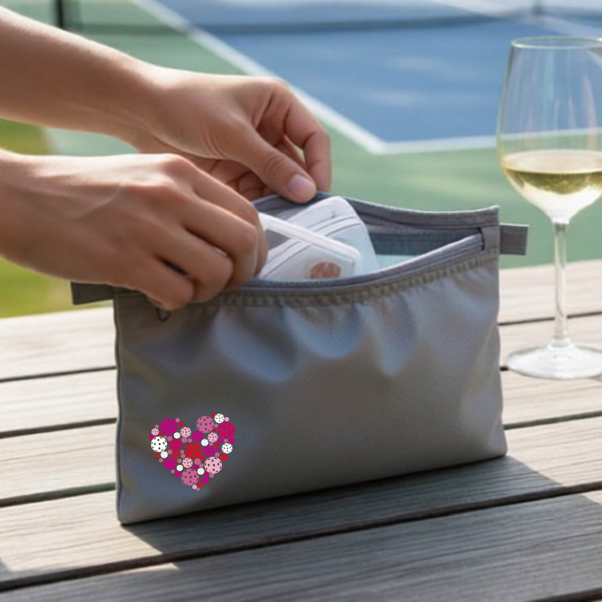 A gray zippered toiletry bag with a heart-shaped pattern in pink, purple, and white, pickleballs shown on a wooden table with a wine glass and a person's hands in the background.
